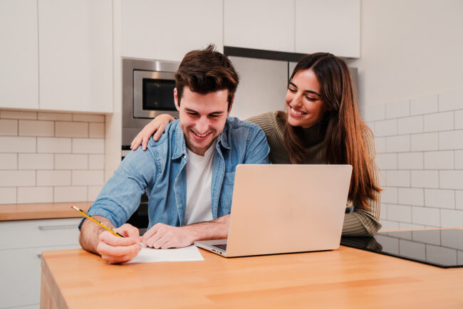 Young couple browsing on internet using a laptop searching a mortgage and calculating their budget sitting at home. Handsome man writing the bank accounts on notebook while his wife supports him. Looking at real estate